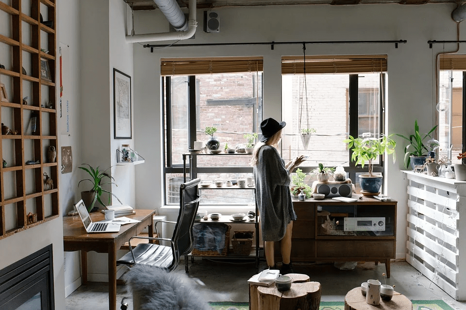 laptop placed on the wooden table, a girl wearing a hat standing near the window, two big windows with blinds