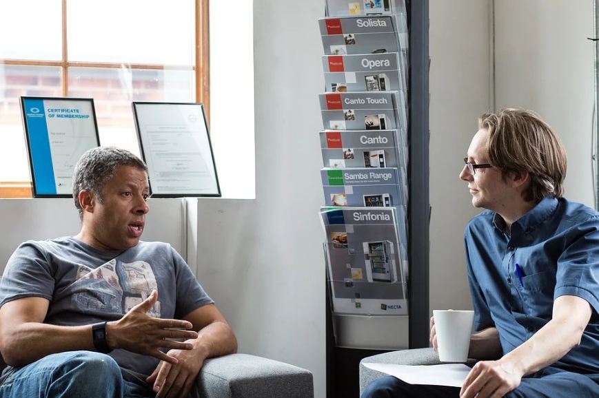 two men in a face-to-face conversation, one is explaining, one is holding a white mug