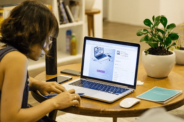 A woman working on her laptop, on a table, with a notebook, and a potted plant