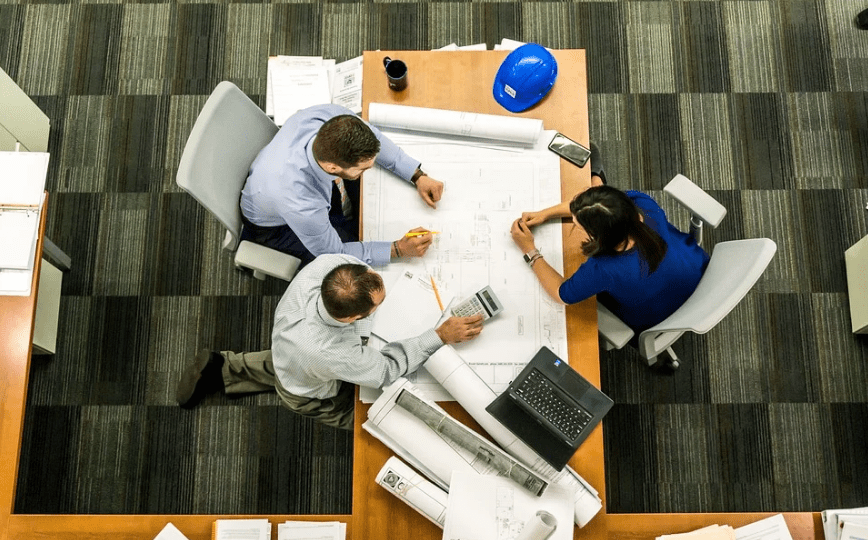 three people discussing, long wooden desk, big and rolled papers on the desk