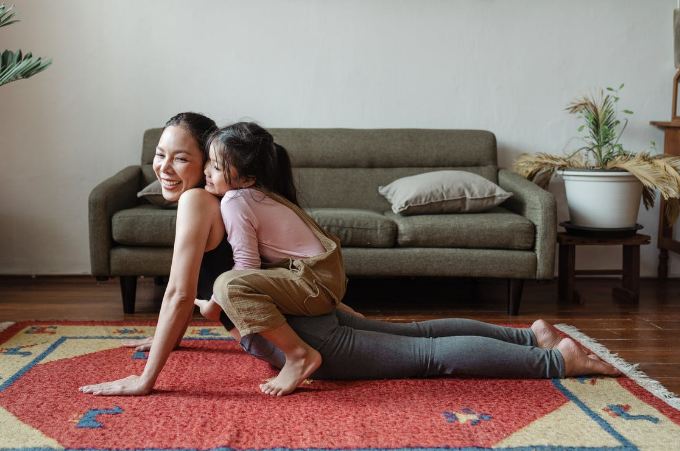 a woman doing yoga with her daughter on her back; there is a sofa, a table, and a plant in the background