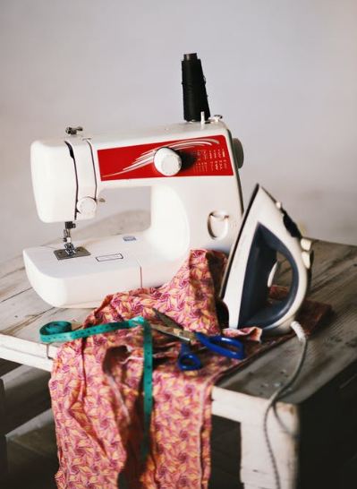 An Image Displaying A White Sewing Machine, Clothes, Iron and Scissor.