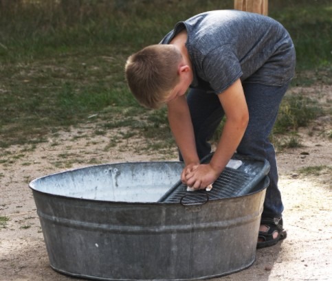 A boy washing clothes in a metal tub using a washboard