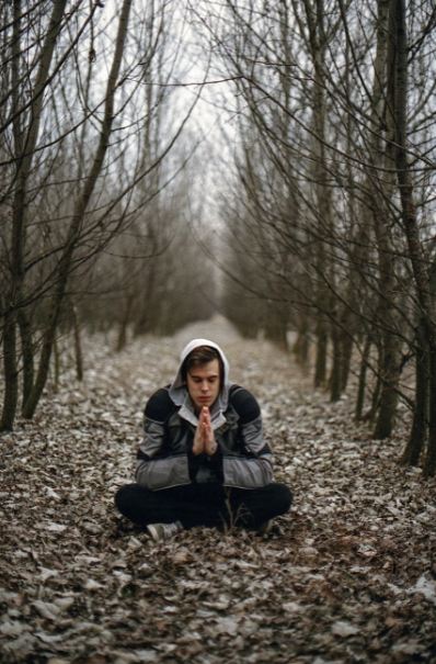 man-wearing-gray-black-zip-hoodie-jacket-praying-in-between-black-tree-during-daytime