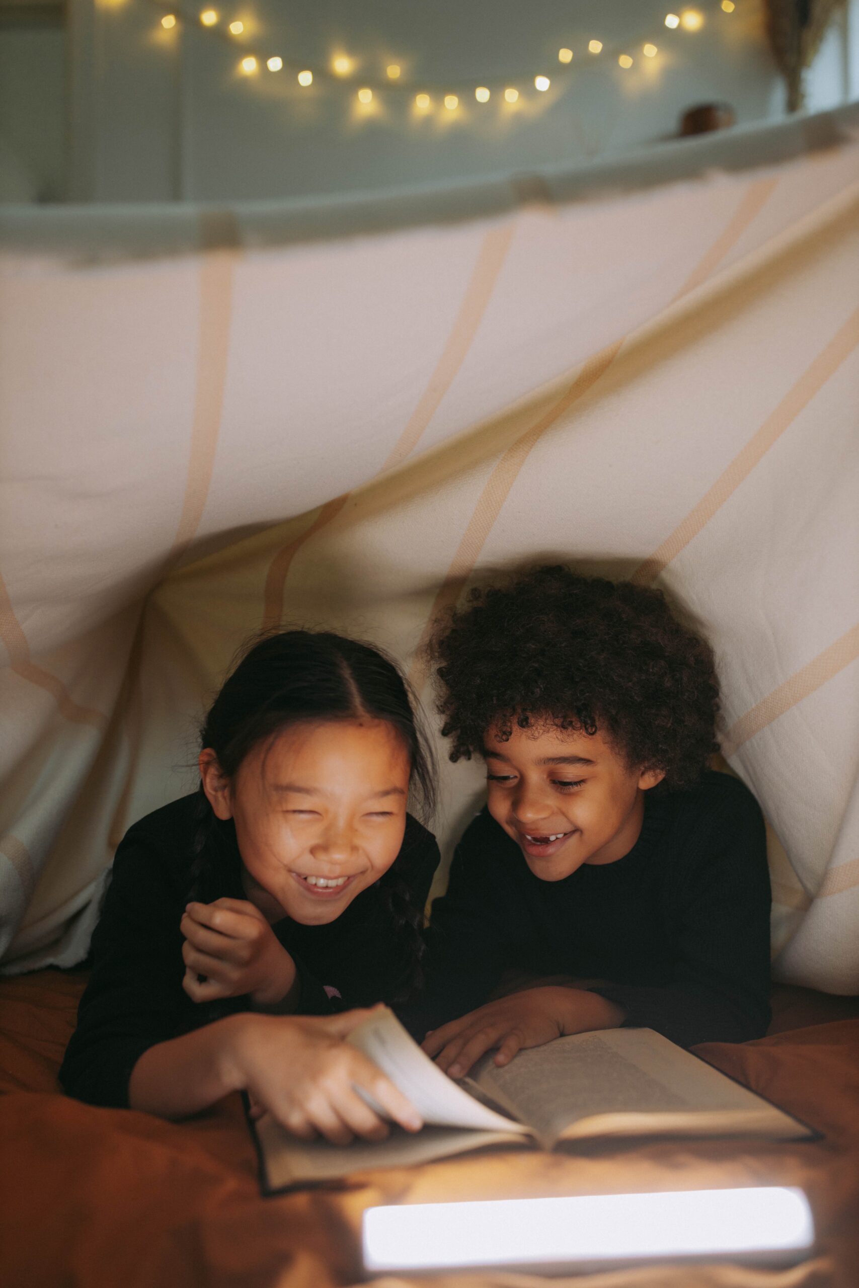 a young girl and boy reading a book while under the blanket